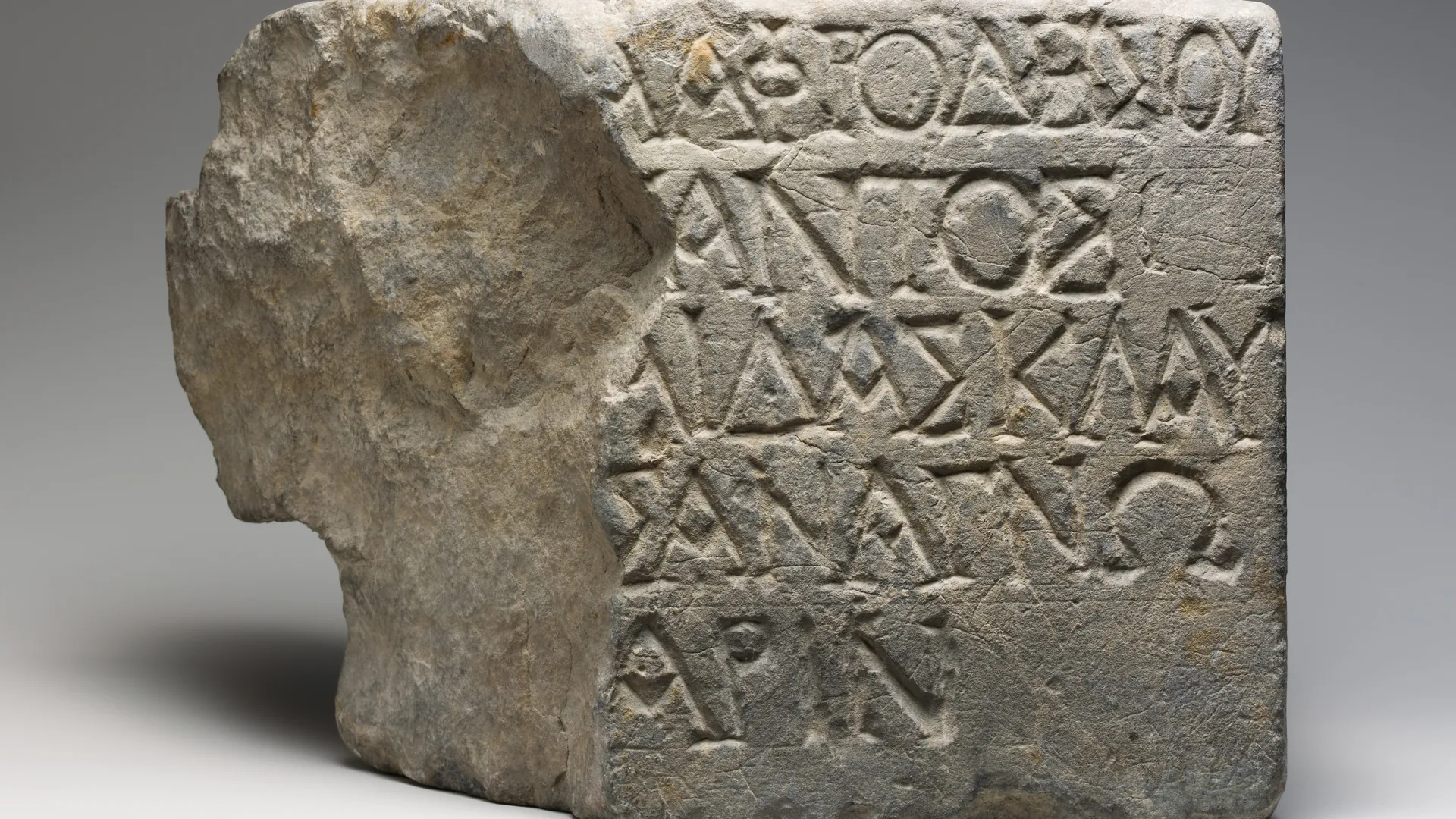 A fragment of a carved Roman marble inscription, five lines of Greek capitals cut into the surviving face, the left side broken away.